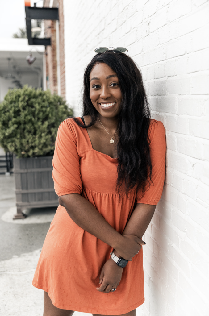 Jordan Chapman - Team Orders About Us Photo; young black woman in an orange dress with sunglasses on her head, arms crossed in front while smiling