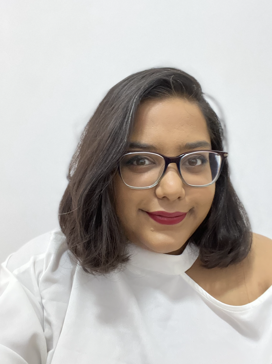 Fadia Ramjuan - Team Orders About Us, a brown skinned woman with classes, with red lipstick and a white top in front of a cream background with a subtle smile